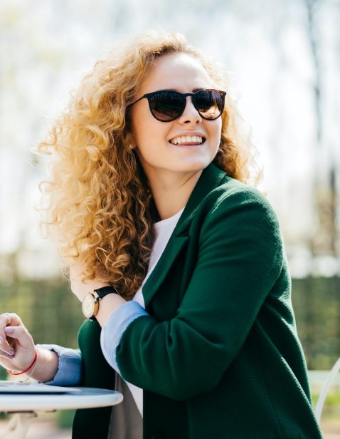 Happy stylish woman with curly light hair wearing sunglasses working with laptop outside in park